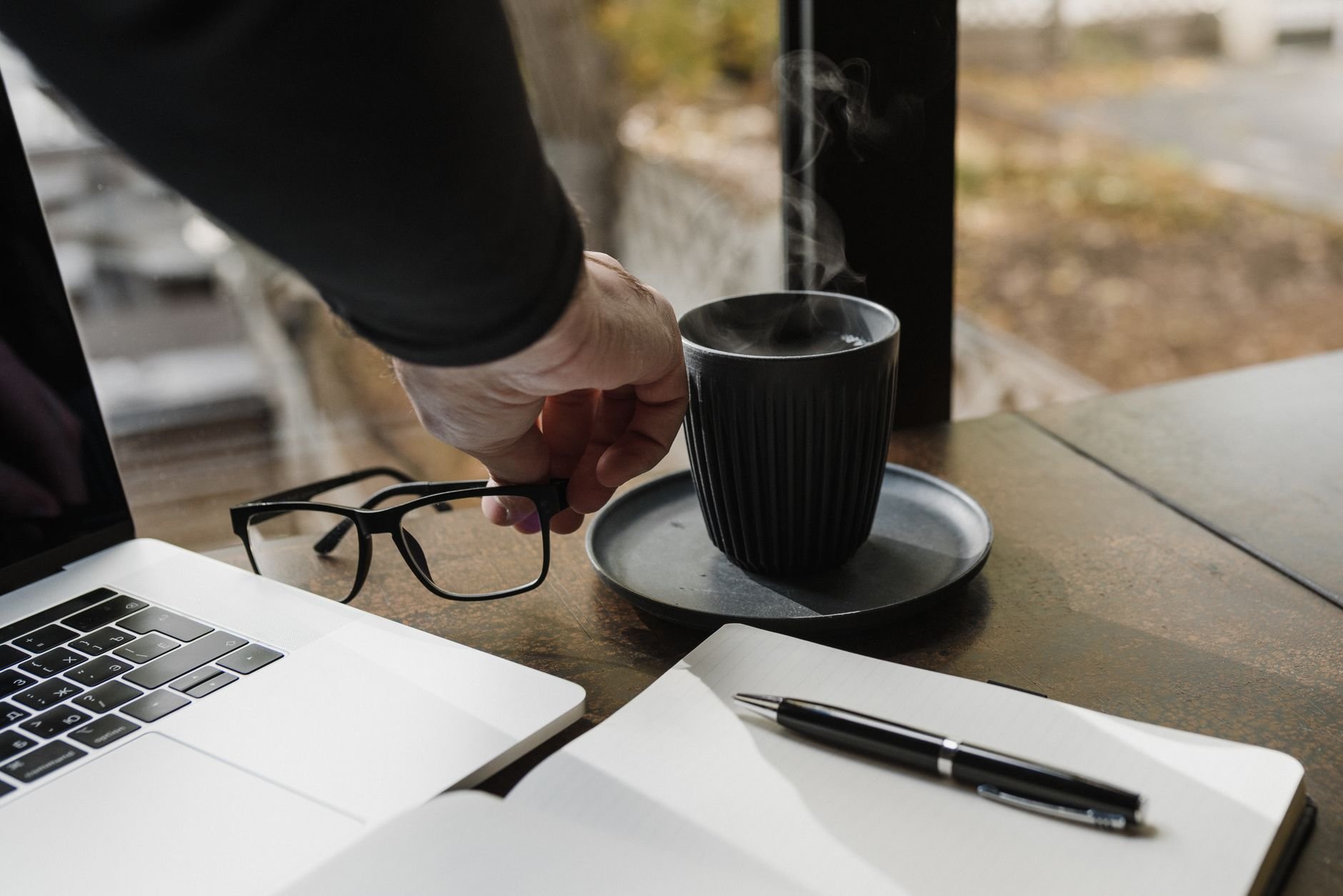 A hand reaches for a steaming cup of coffee near a laptop and open notebook, creating a cozy workspace vibe.