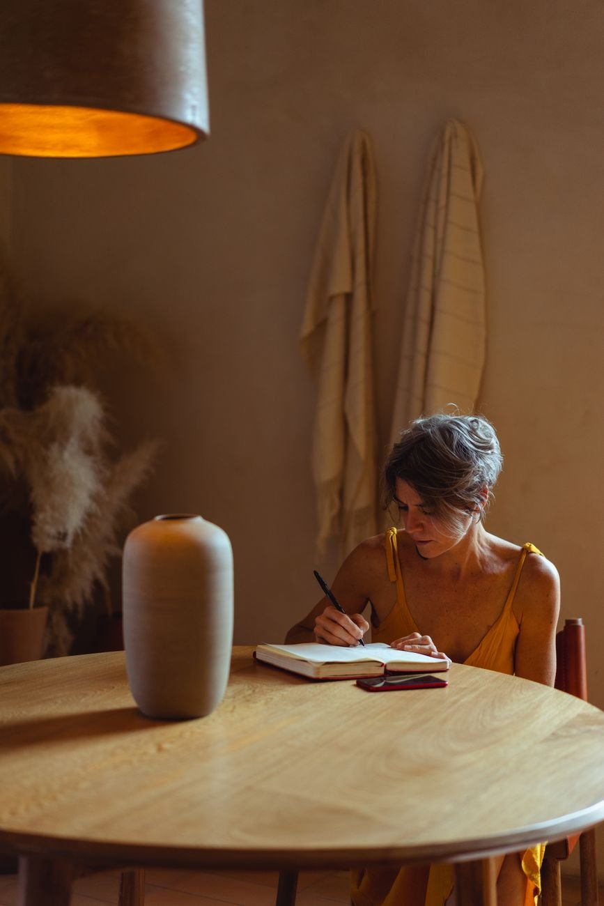 Elderly woman writing in a notebook at a warm, cozy home office with ambient lighting.