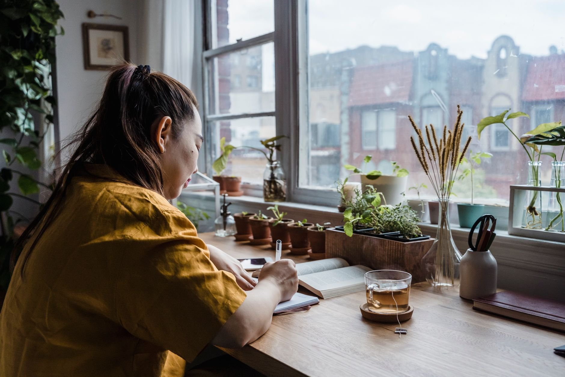 Asian woman writing in notebook in a cozy home office by window with plants.